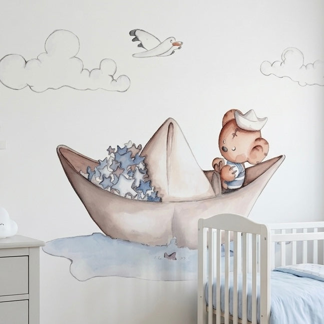 Vinilo decorativo infantil que muestra un oso marinero en un barquito de papel, rodeado de nubes y una gaviota.