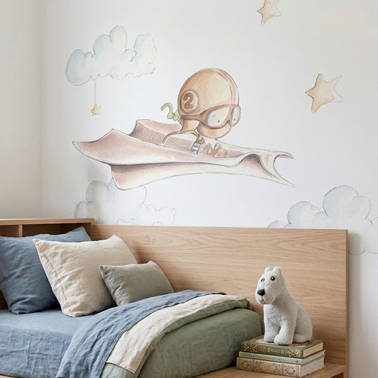 Vinilo decorativo infantil de un niño volando en un avión de papel, rodeado de nubes y estrellas en tonos suaves.