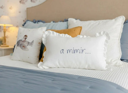 White embroidered children's pillow with ruffled edges and 'a mimir...' text on a cozy bed.